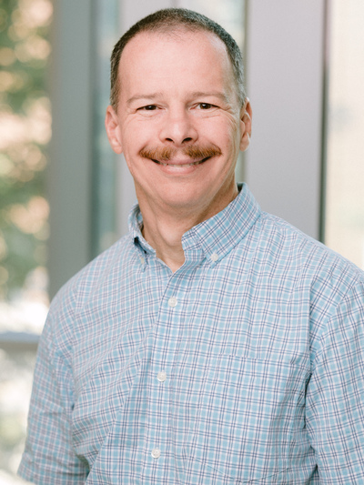 Portrait photograph of Yuri Kovchegov, Professor of Physics, smiling in a light blue checkered shirt